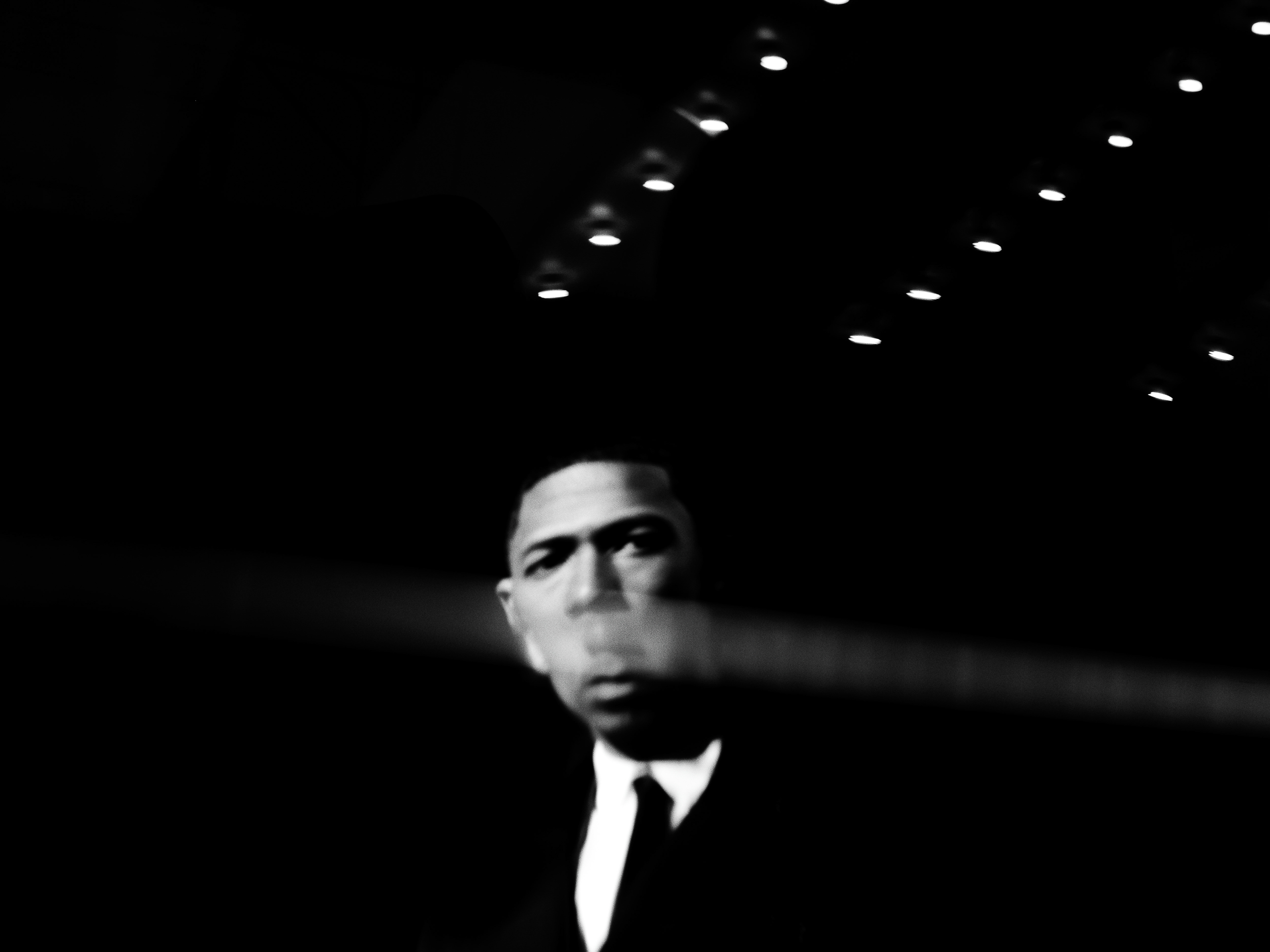 Black and white portrait of a man in a suit and tie standing indoors, lit by spotlights against a dark background.