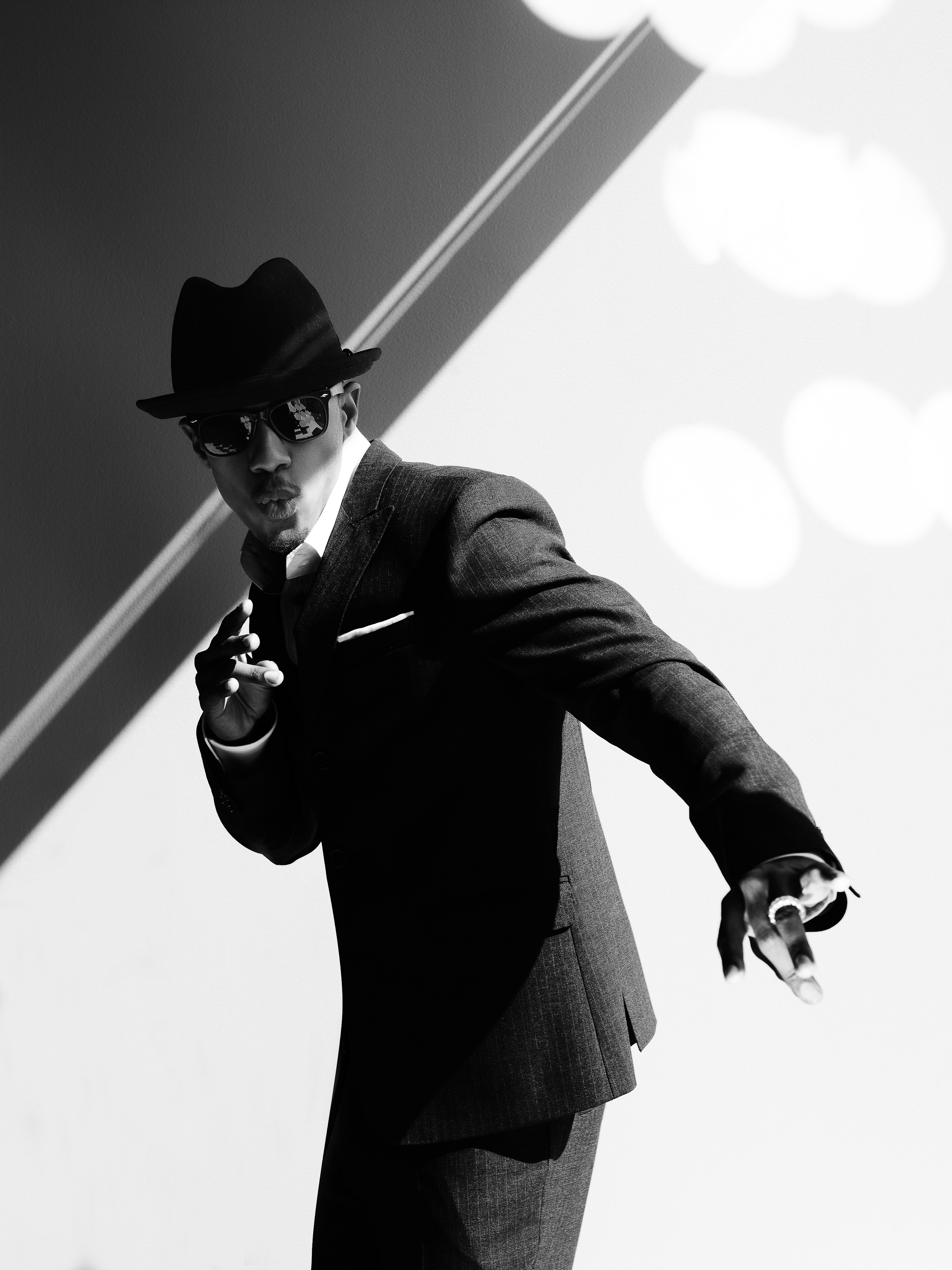 Black-and-white portrait of a stylish man wearing a suit, fedora, and sunglasses, in a dynamic pose against a geometric background.