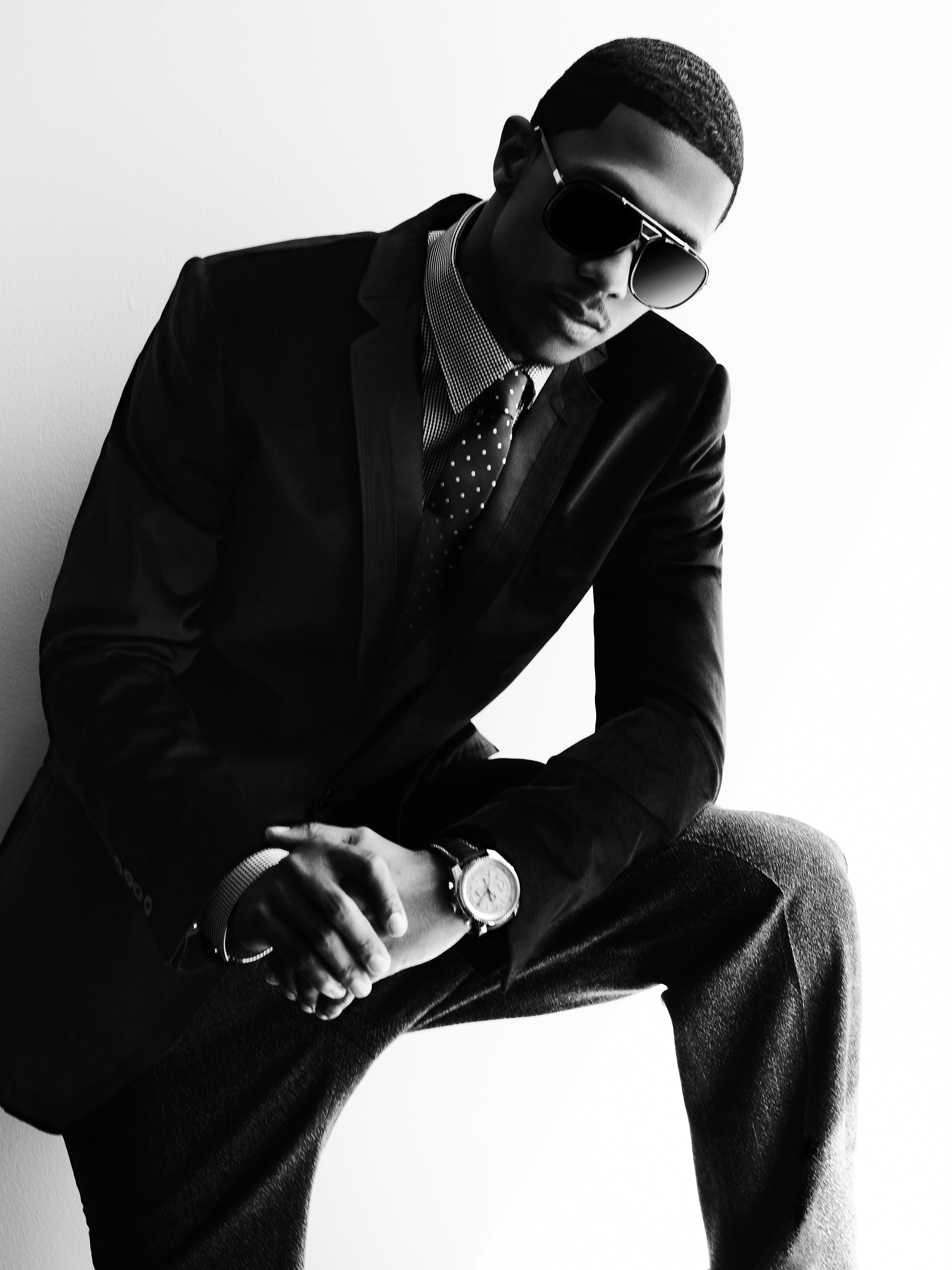 Black and white studio portrait of an African American male model in a dark suit and sunglasses, posing with his hands clasped on his knee.