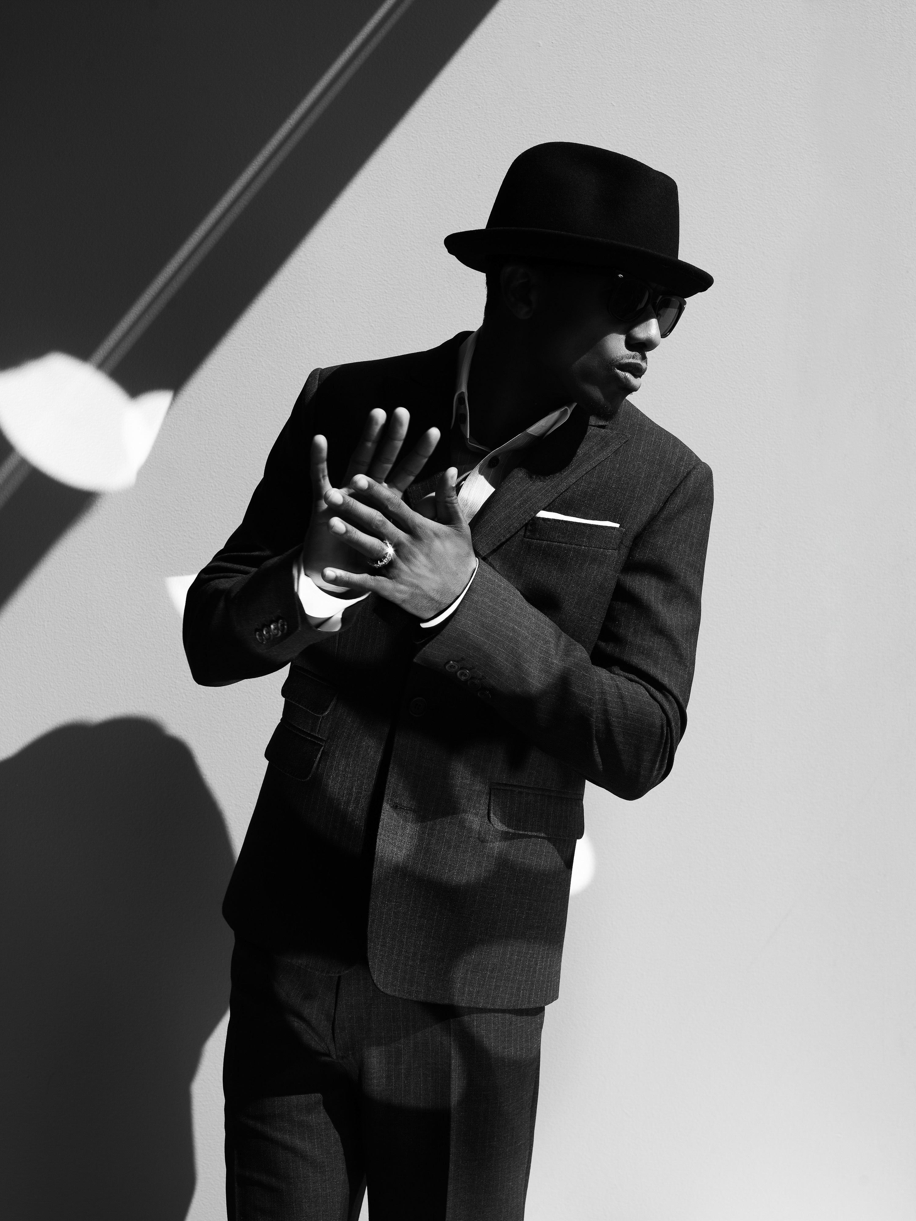 Black and white portrait of a stylish adult male wearing a hat and suit with patterned shadows in an indoor setting.