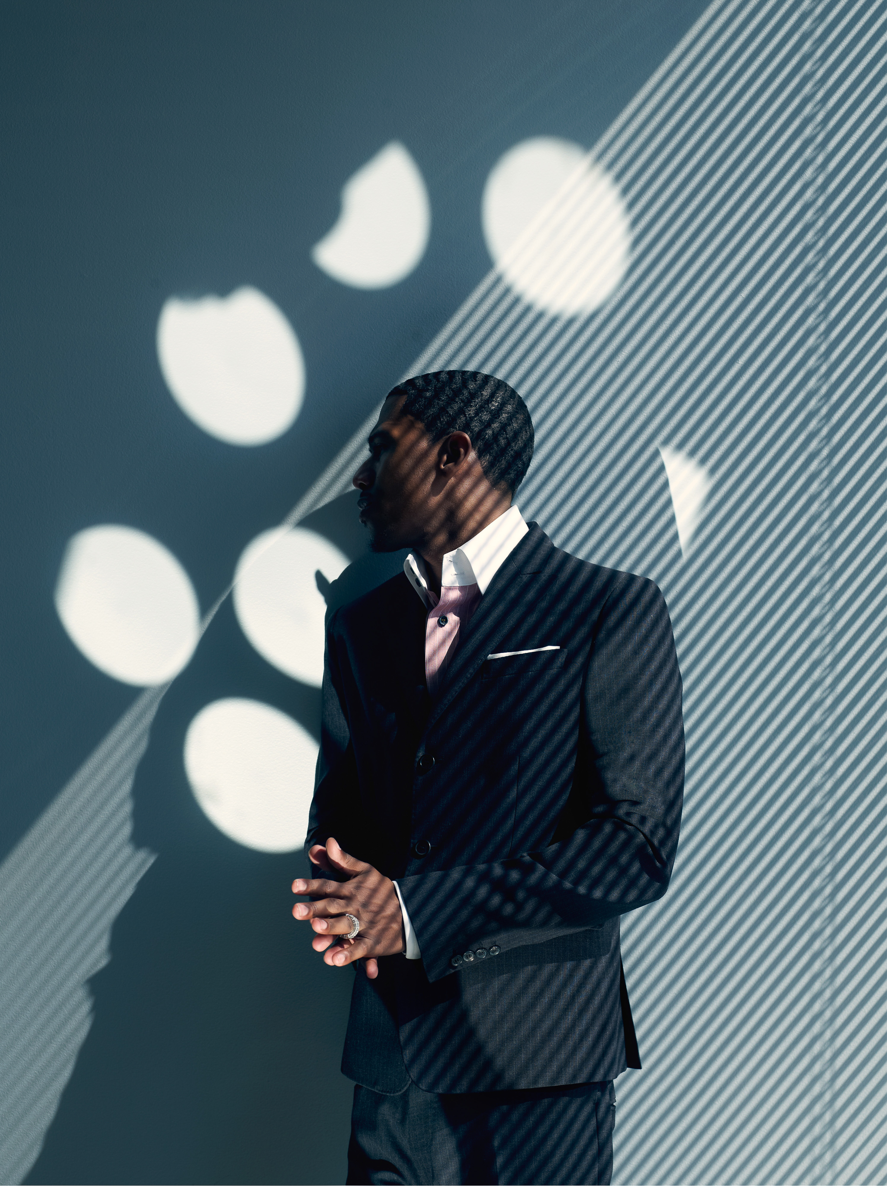 Studio portrait of Black man in dark suit and pink shirt standing against grey wall with linear and circular light patterns.