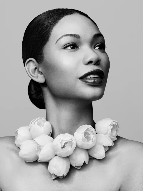 Monochrome portrait of a smiling Black woman wearing a floral collar of white tulips against a neutral grey studio backdrop.