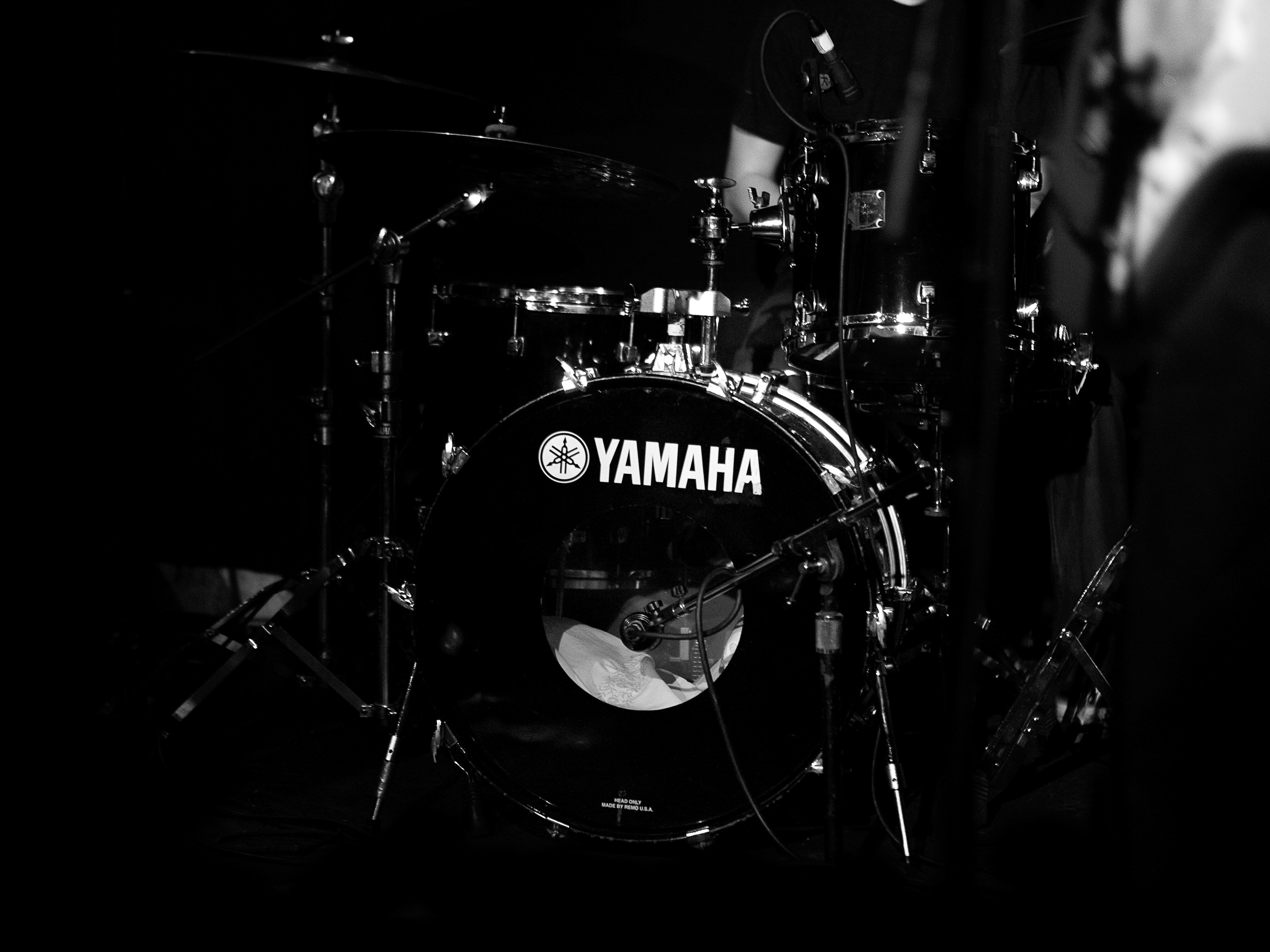 A black and white shot focuses on a Yamaha drum set with a visible logo, featuring cymbals, drum hardware, and a darkened background.