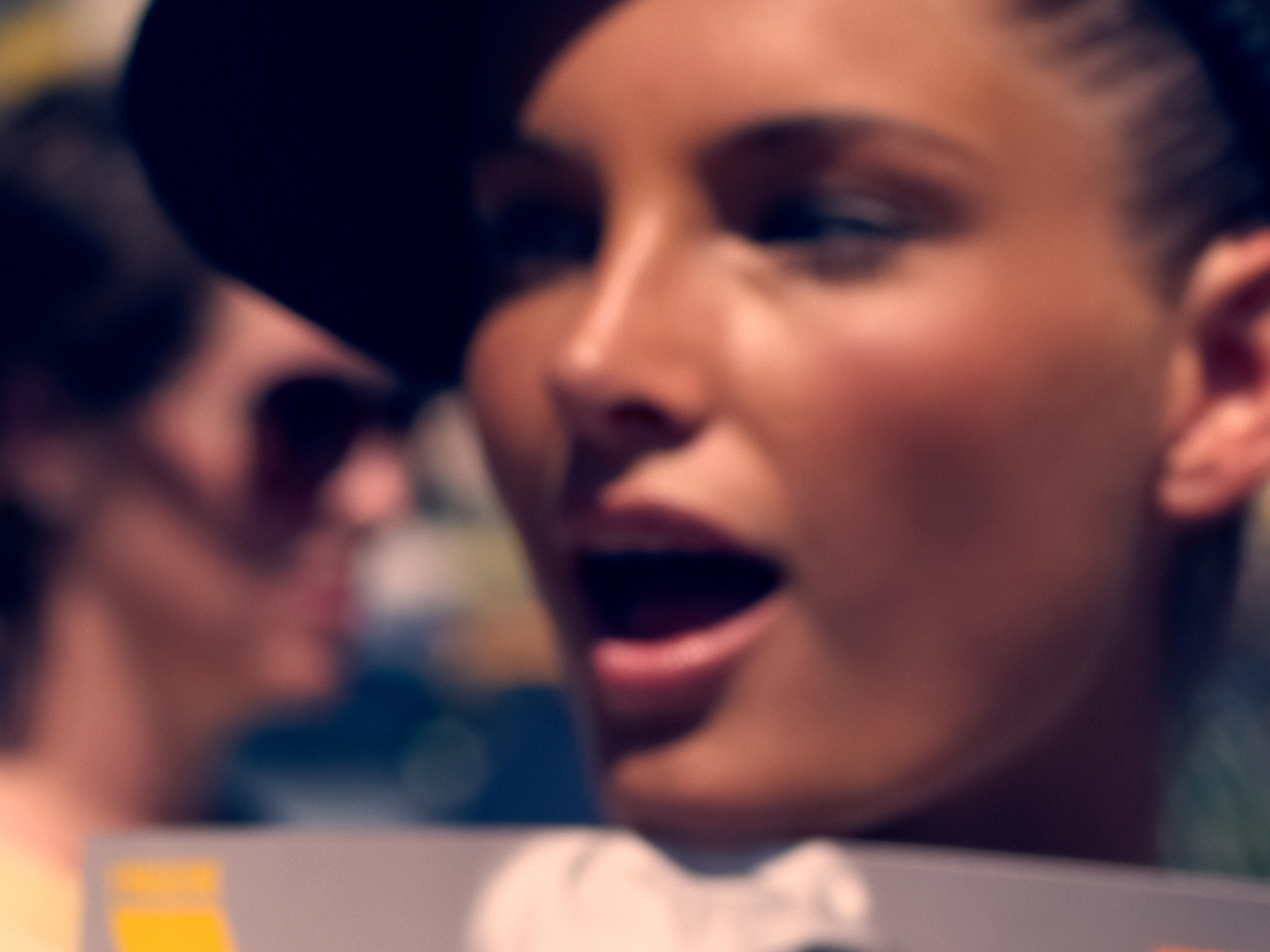 Close-up of a woman with her mouth open, wearing a dark hat, and another person blurred in the background.
