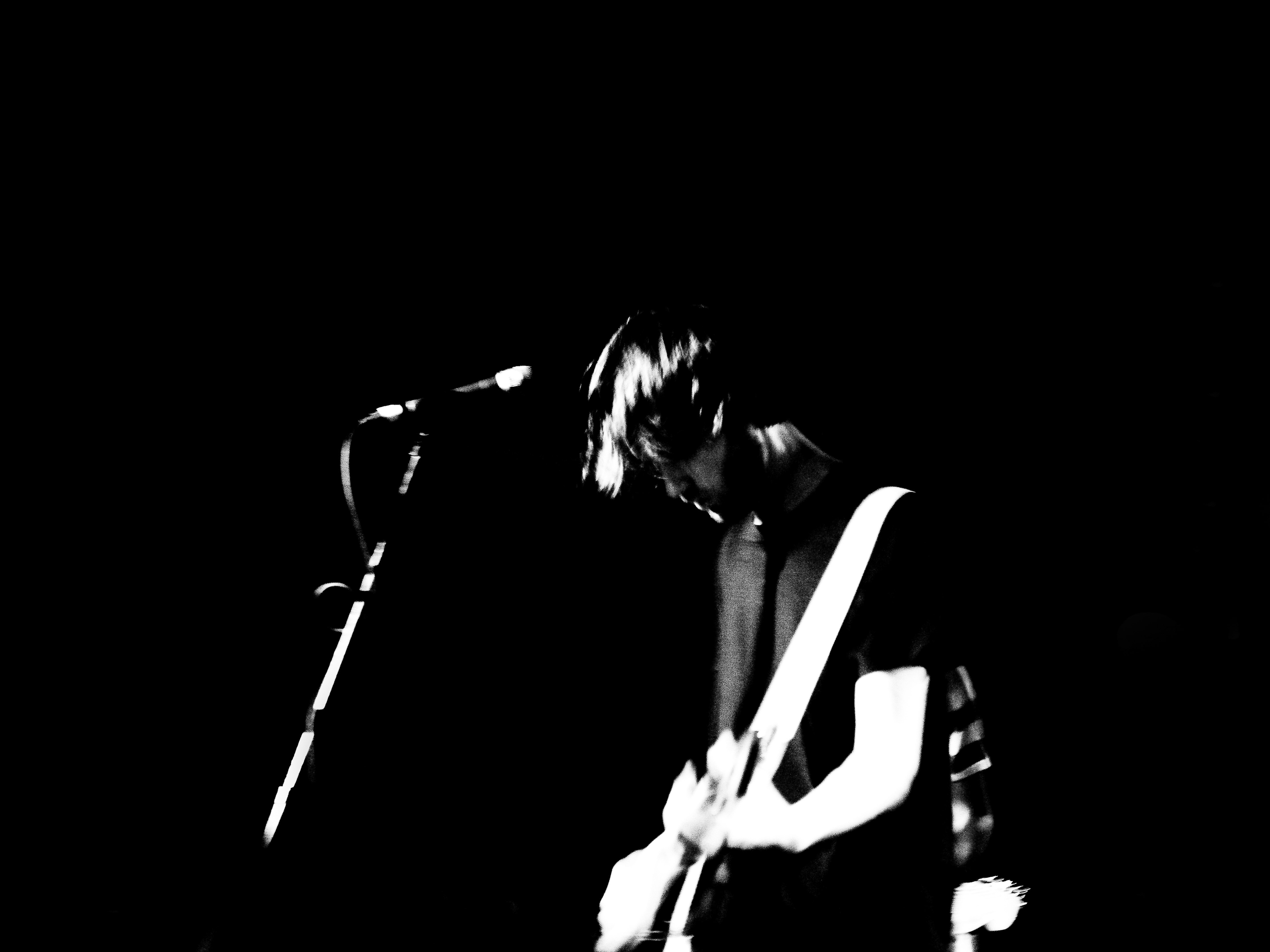 A monochromatic shot captures a musician singing into a microphone and playing an electric guitar during a live music performance.