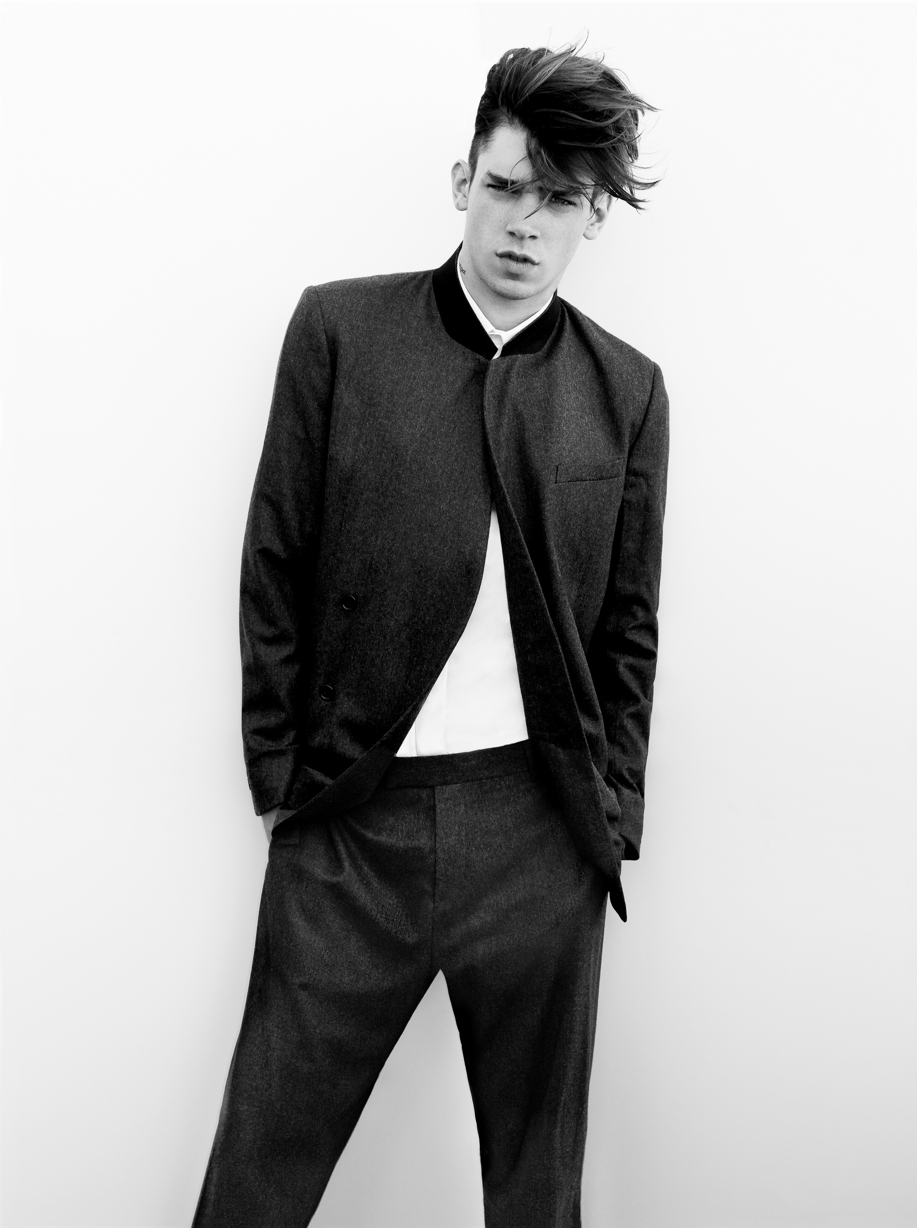 A young man with swept-over hair poses in a dark mandarin-collar suit and white shirt against a white seamless background.