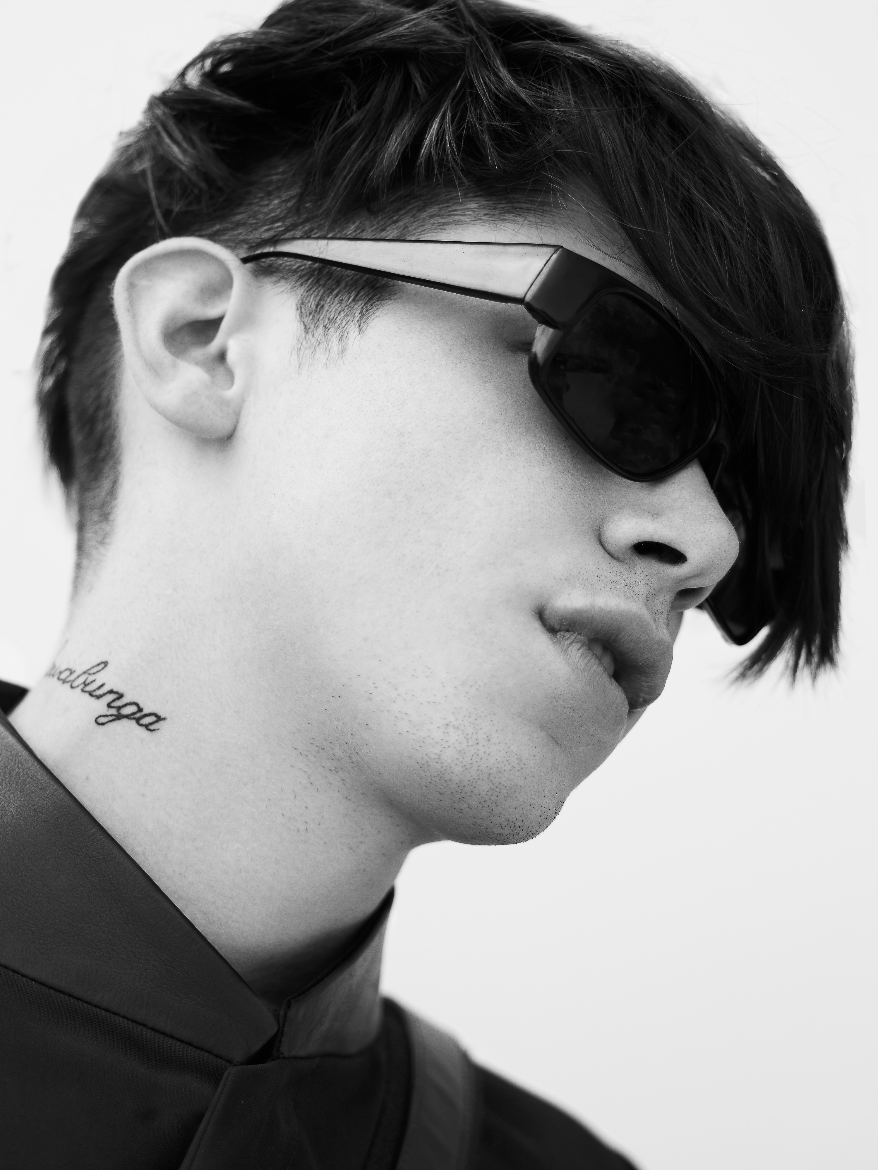Black and white portrait of a young adult male with dark hair, sunglasses, and a visible tattoo on his neck in a studio setting.