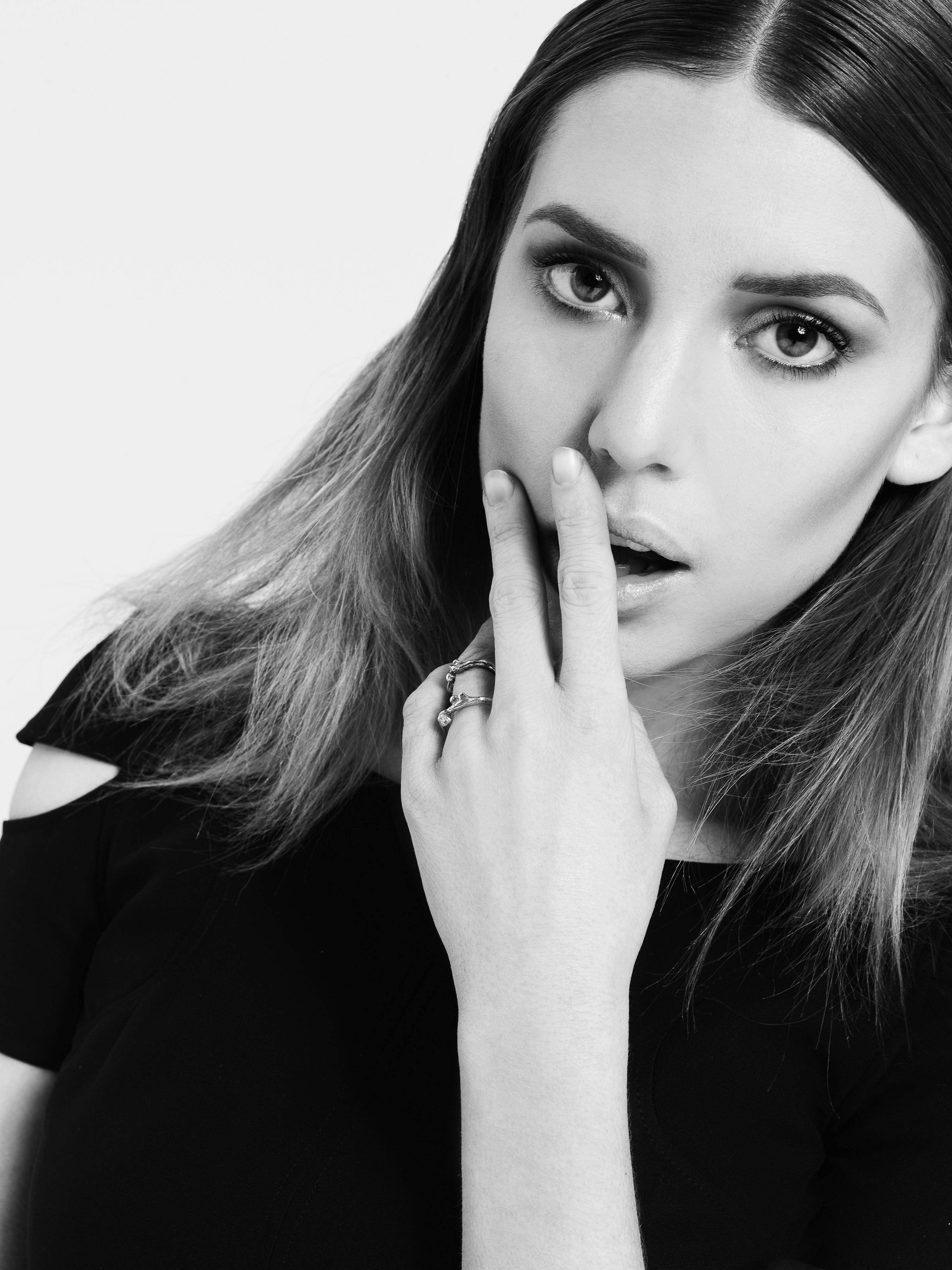 Portrait of an adult woman with long hair, wearing a dark top and ring, with fingers near her mouth, against a white background.