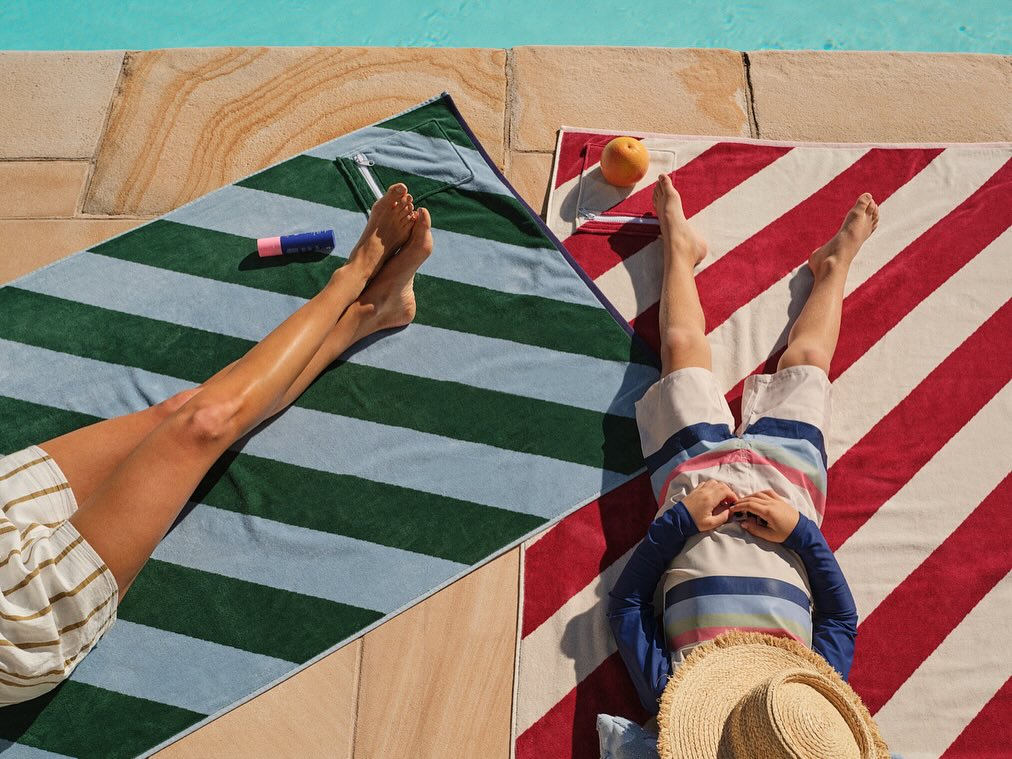 Aerial view of an adult and a child laying on striped towels by a pool with accessories on a sunny day, enjoying summer vibes.