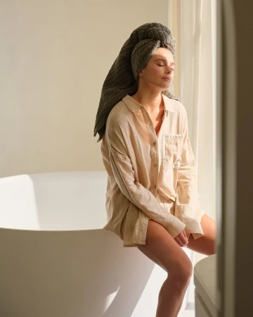 Woman with towel turban sits on white bathtub edge in sunlit room wearing oversized shirt with her eyes closed, enjoying spa relaxation.