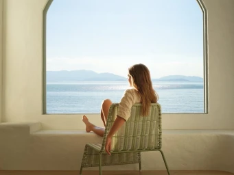 Woman sitting in a wicker armchair looking out a large arched window at the ocean for Adairs Spring 2024 campaign