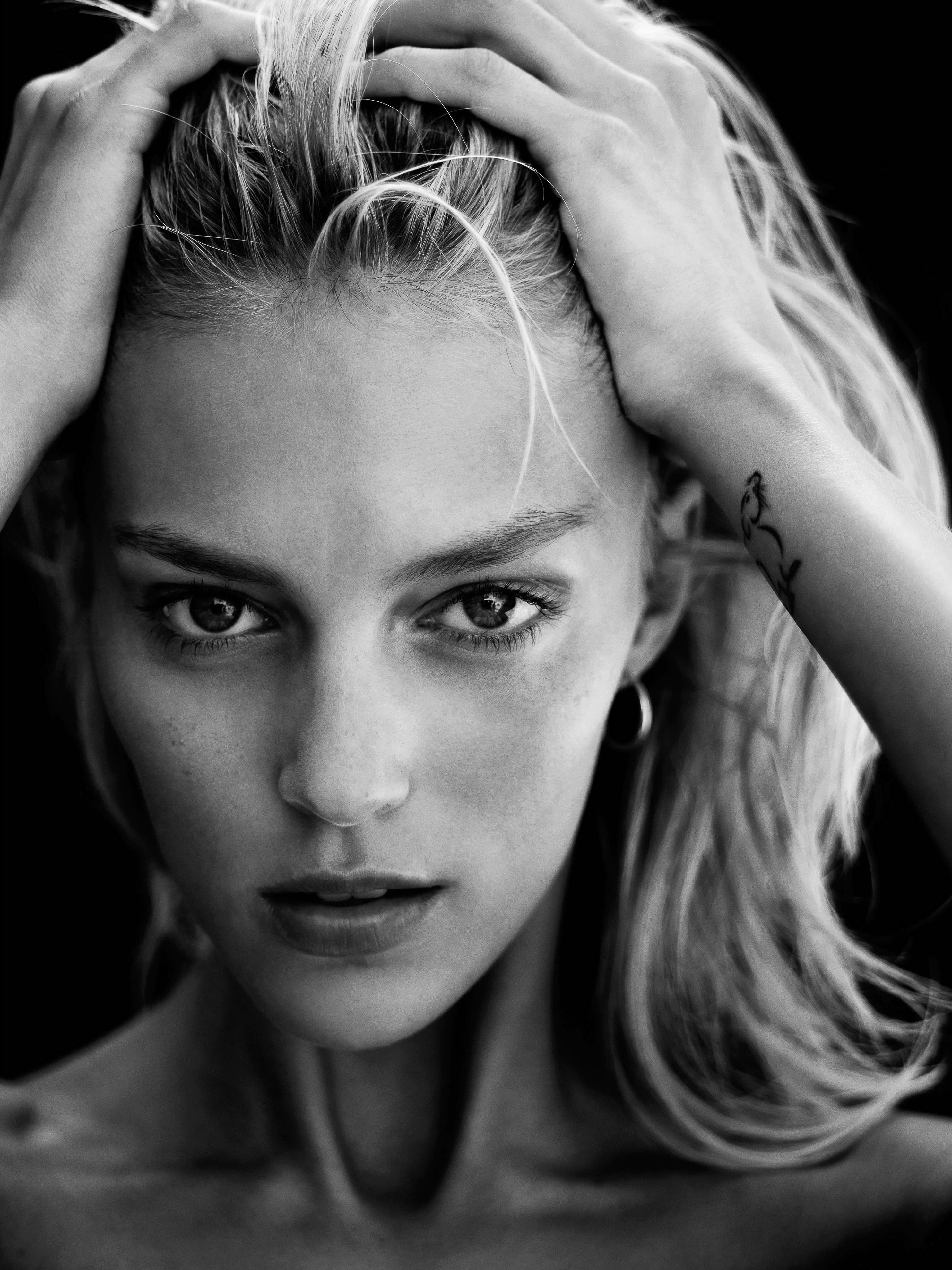 Close-up black and white portrait of an adult woman with blonde hair, hands in her hair, hoop earrings, and a black backdrop.