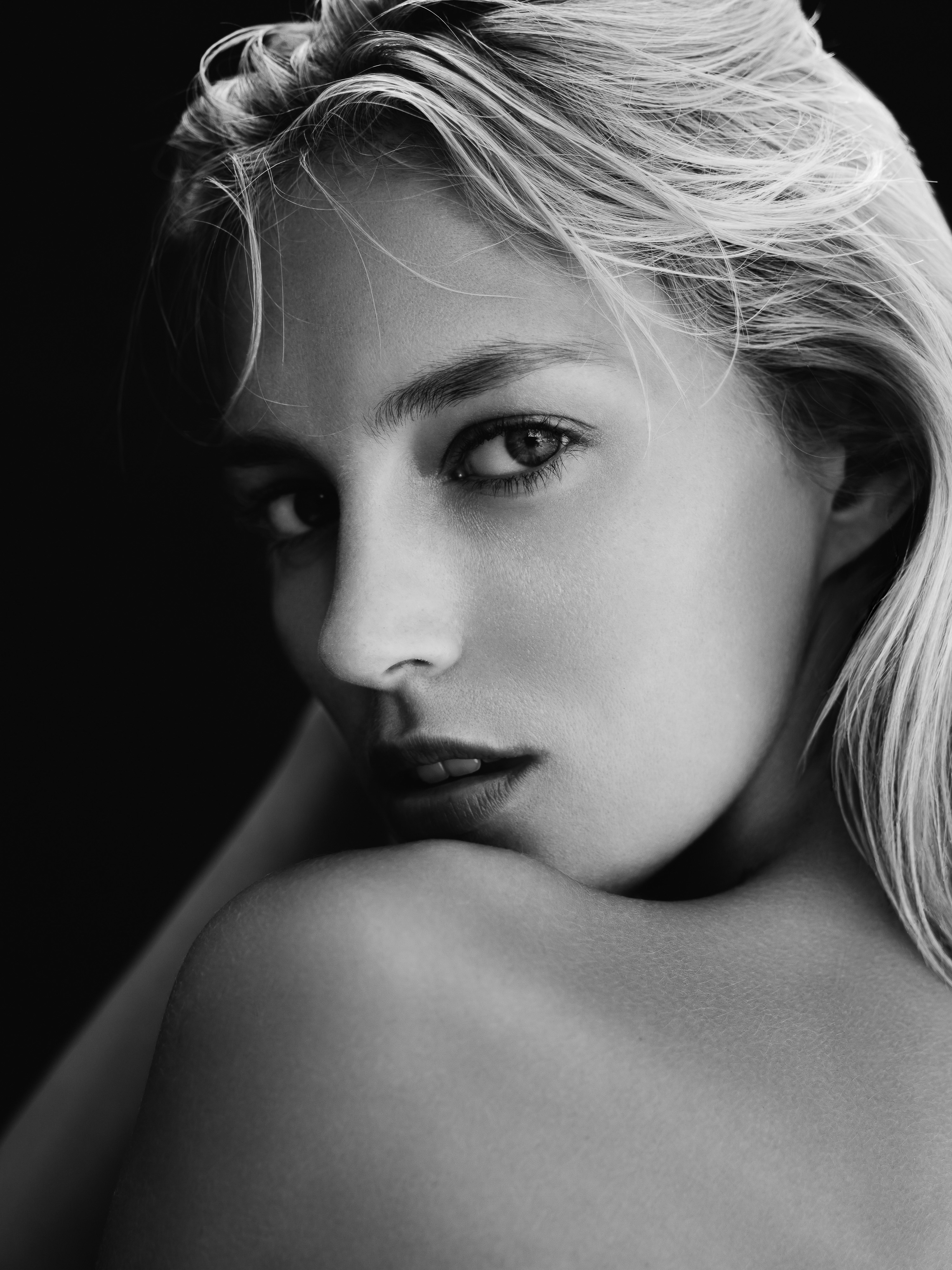 Black and white portrait of a blonde woman with bare shoulders, looking at the camera in a dimly lit studio.