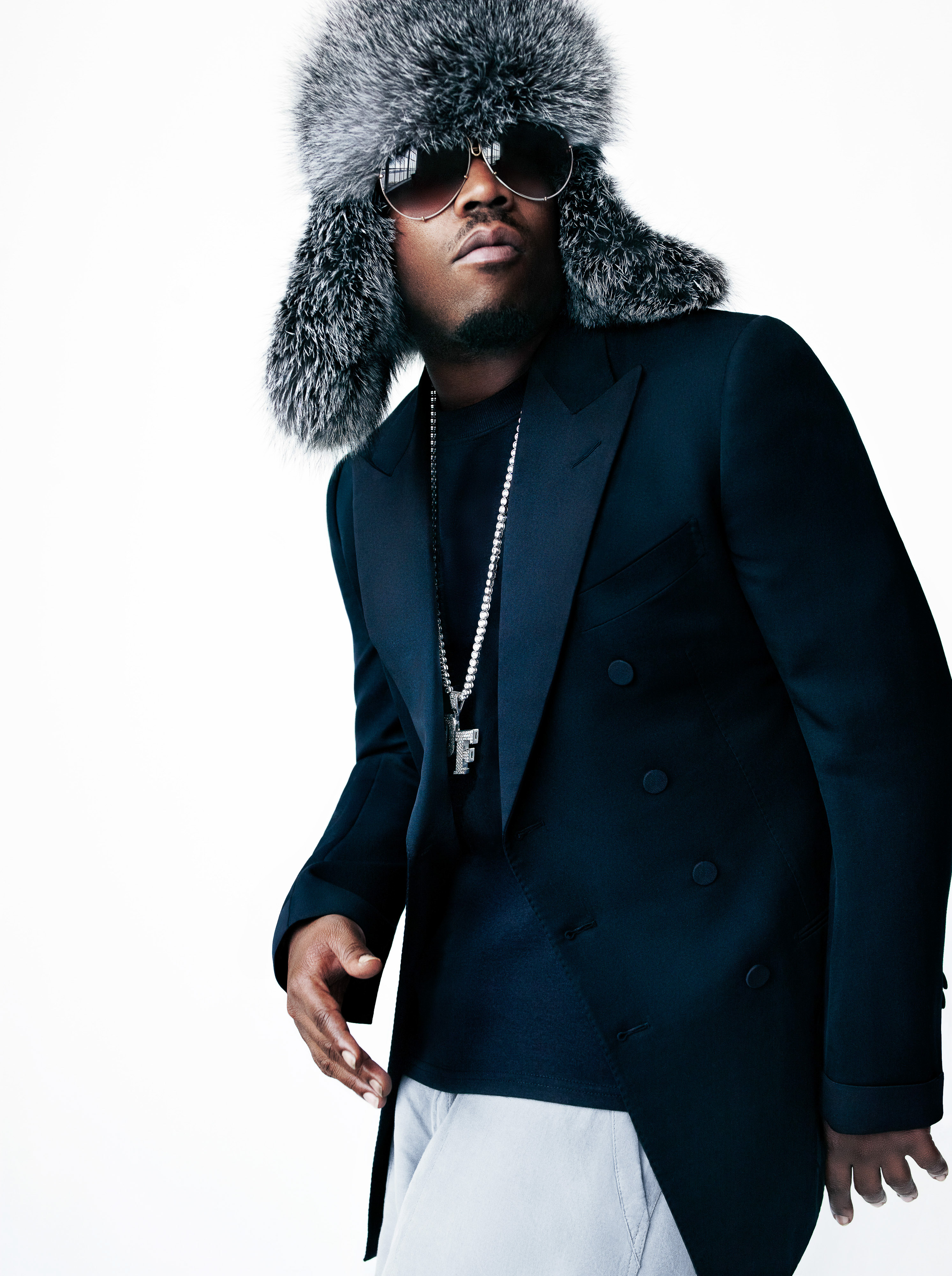 Adult male wearing a fur hat, blazer, chain, and sunglasses poses against a white background in a studio portrait.