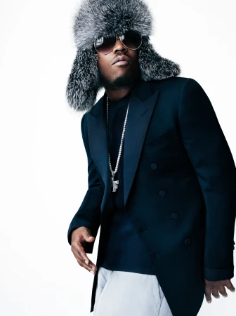 Adult male wearing a fur hat, blazer, chain, and sunglasses poses against a white background in a studio portrait.