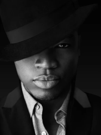 Black and white portrait of a handsome adult male with a hat, wearing a dress shirt and suit jacket, posing against a dark background.