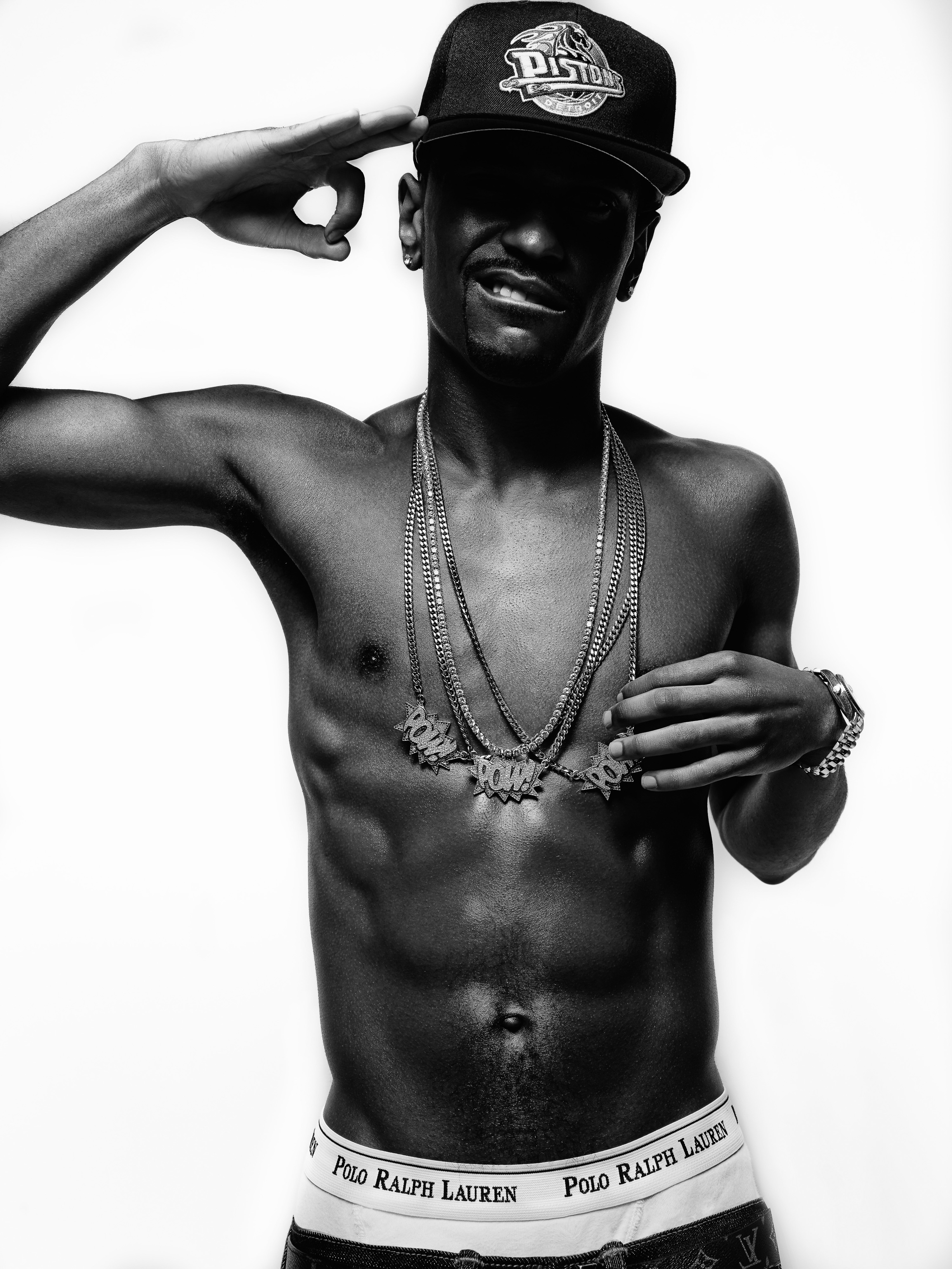 Black and white shot of a shirtless male adult wearing a baseball cap and gold chains posing in front of a white backdrop.