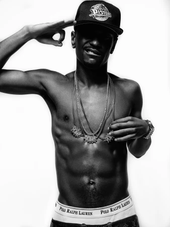 Black and white shot of a shirtless male adult wearing a baseball cap and gold chains posing in front of a white backdrop.