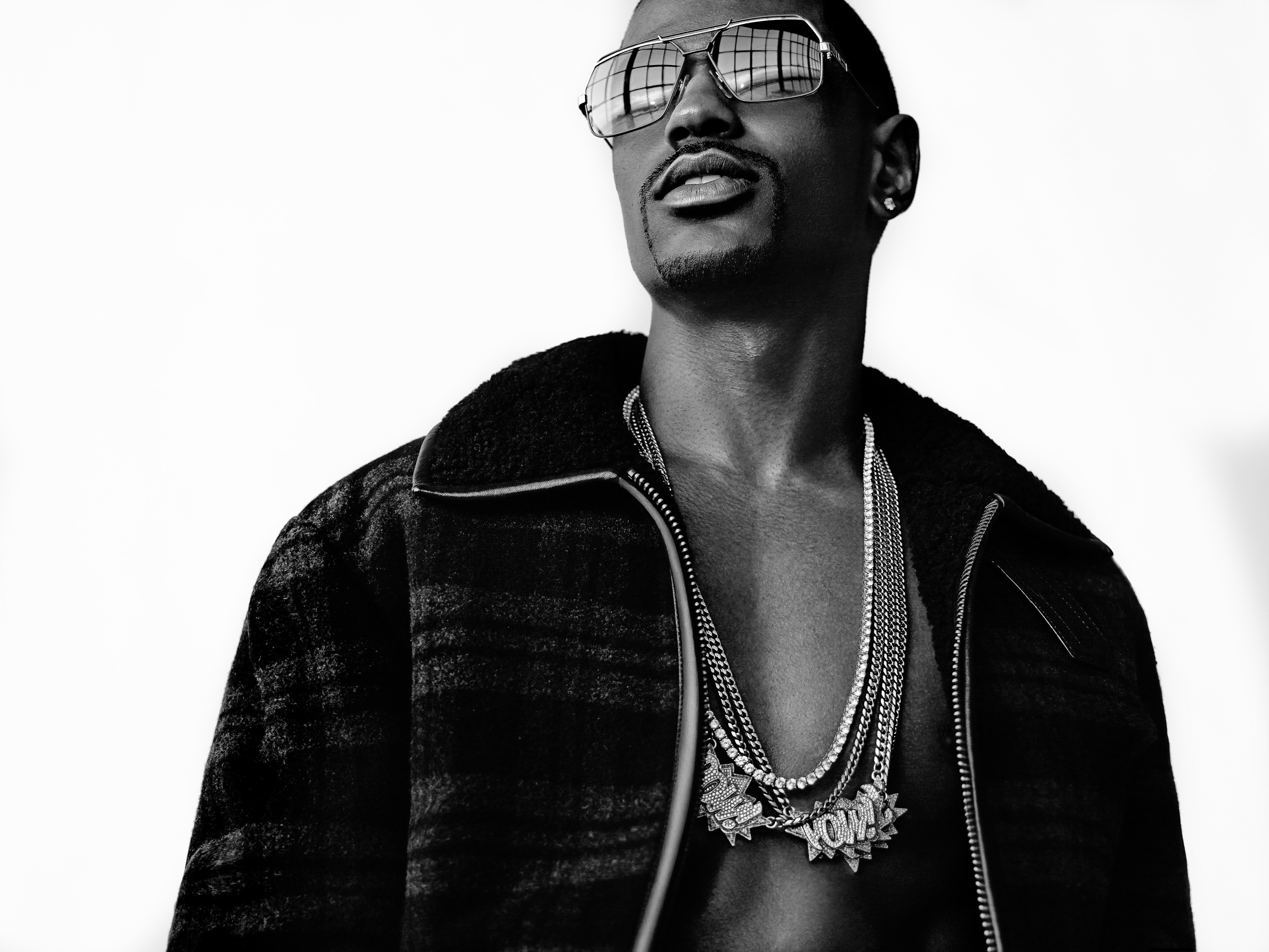 Black and white portrait of a man with sunglasses and layered necklaces, wearing a patterned jacket against a bright background.