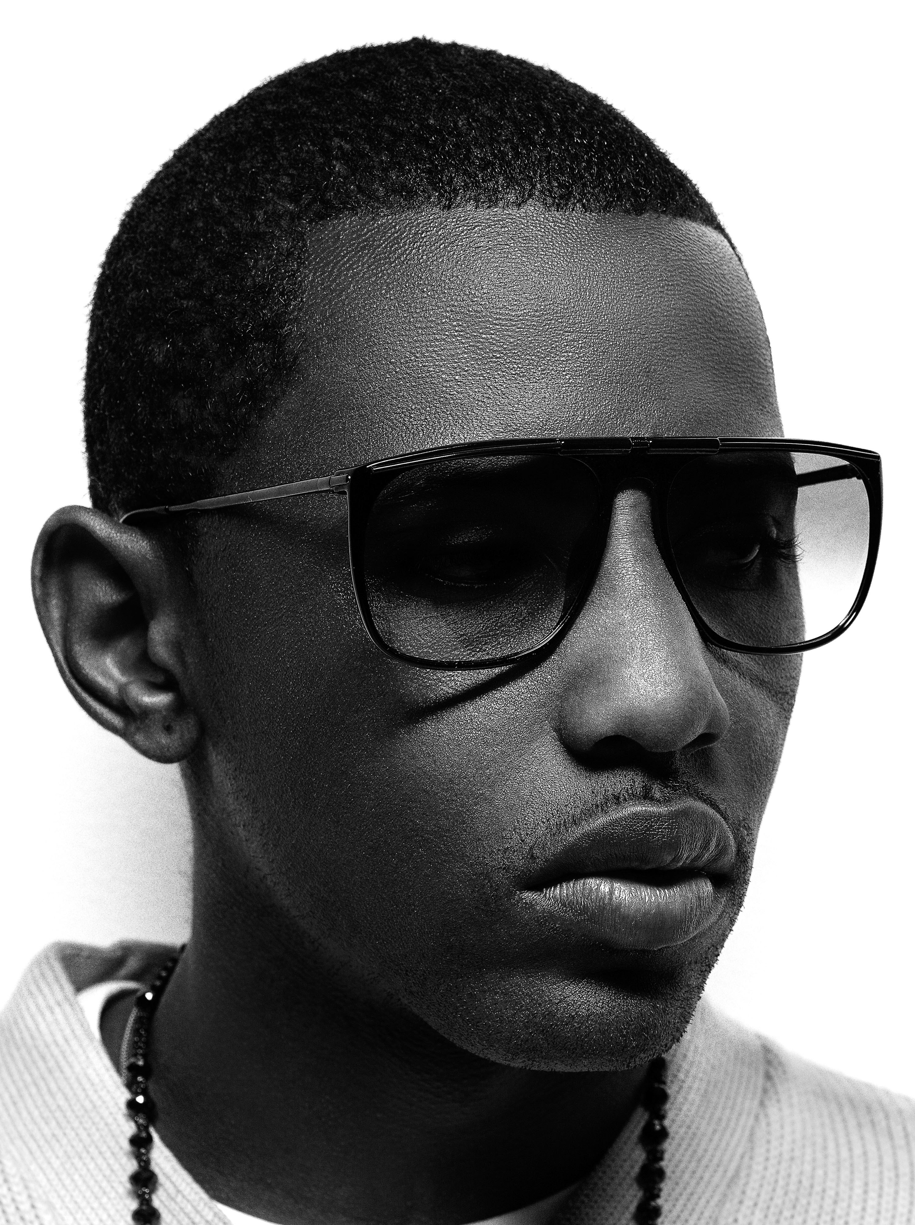 Black and white portrait of a dark-skinned man with close-cropped hair wearing dark sunglasses and a beaded necklace against a white backdrop.