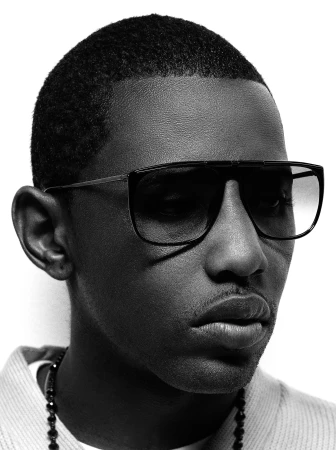 Black and white portrait of a dark-skinned man with close-cropped hair wearing dark sunglasses and a beaded necklace against a white backdrop.