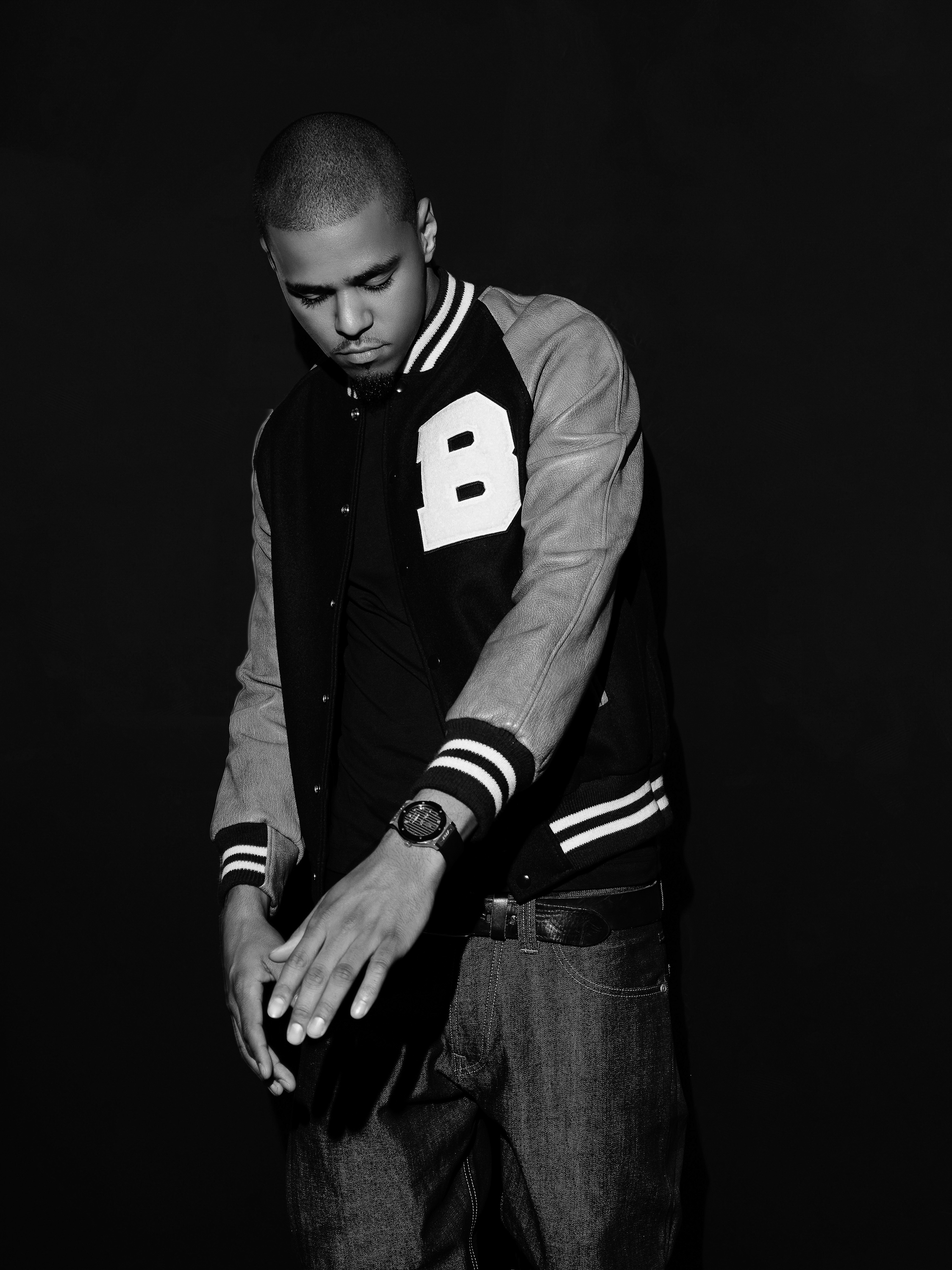 Black and white portrait of adult male in a letterman jacket and jeans against a dark backdrop, looking down with hands clasped.