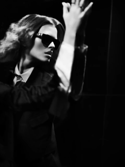 Monochrome portrait of a woman with wavy hair and sunglasses, wearing a suit, against a dark background in a studio setting.