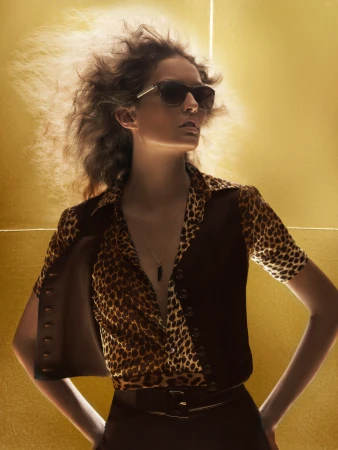A woman with curly hair poses in sunglasses wearing an animal-print blouse and vest in front of a textured gold background.