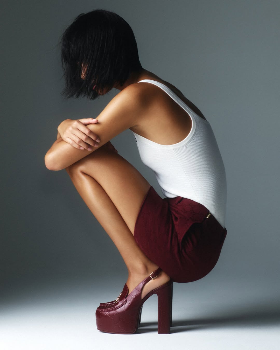 Woman crouches in a studio wearing a white tank top, maroon skirt, and high-heeled shoes with platform soles, lit in a studio environment.
