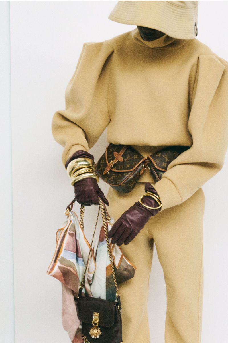 Fashionable person with handbags, wearing a beige outfit, bucket hat, gold bangles and gloves, standing in a bright studio setting.