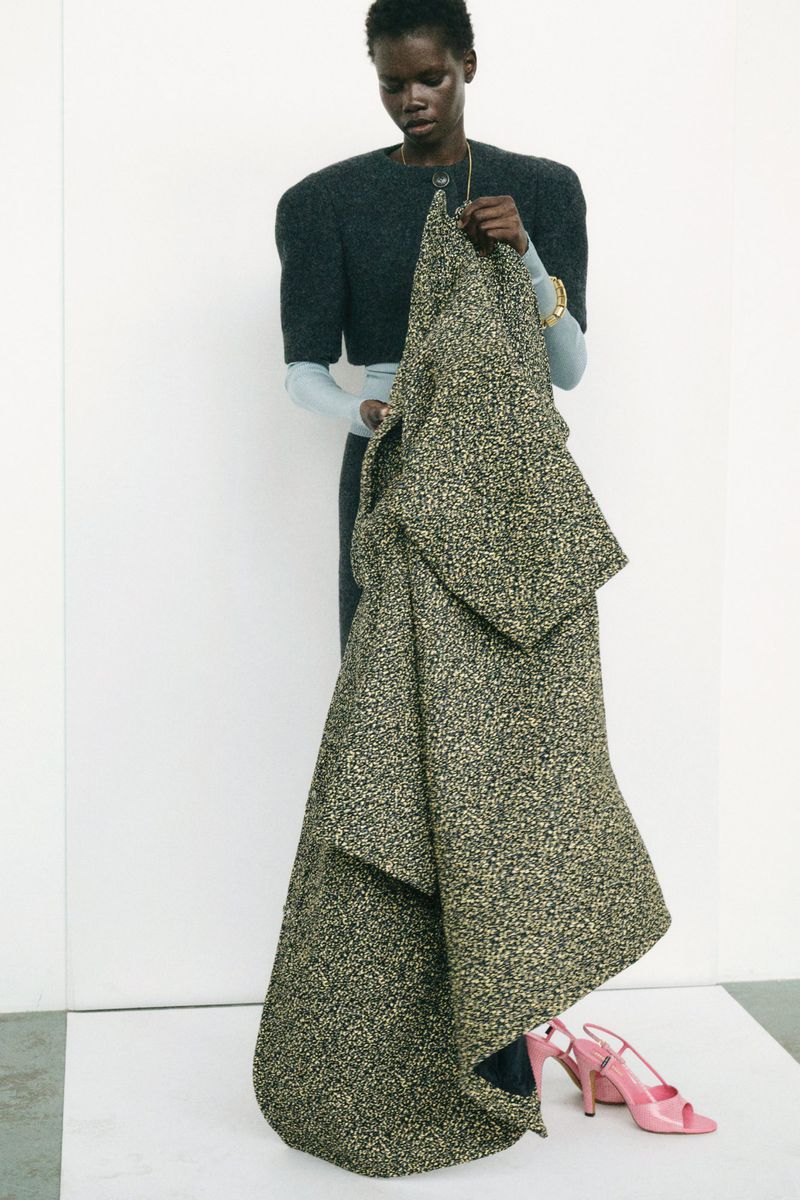 Black female model poses in a photo studio, wearing a structured gray dress and holding a patterned coat with pink high heels on the floor.