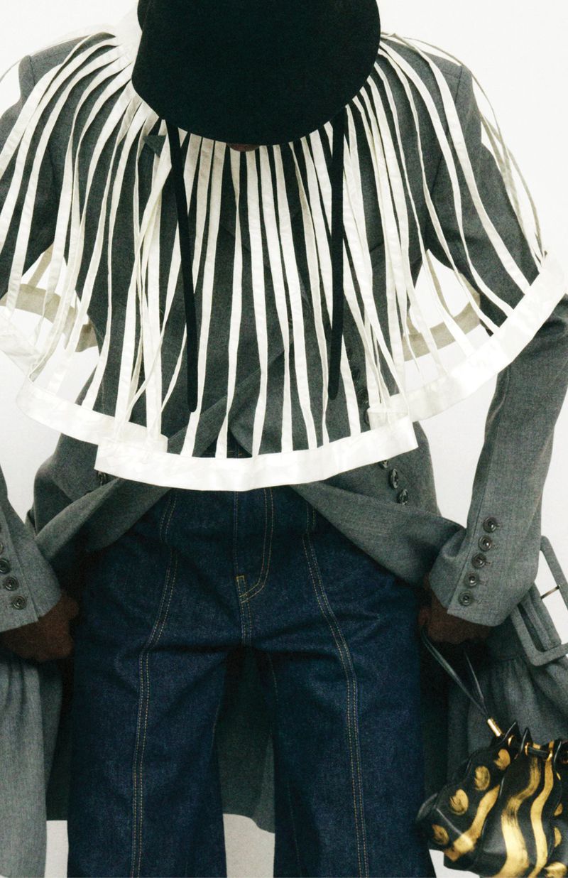 Fashion shot of a human wearing denim jeans, a grey coat and a striped shirt, along with a dark bag, against a white backdrop.