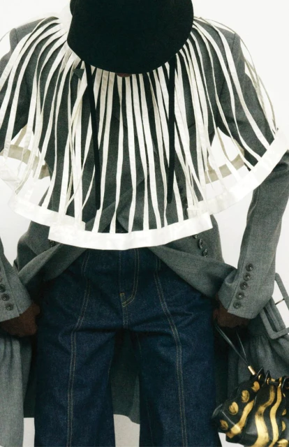 Fashion shot of a human wearing denim jeans, a grey coat and a striped shirt, along with a dark bag, against a white backdrop.