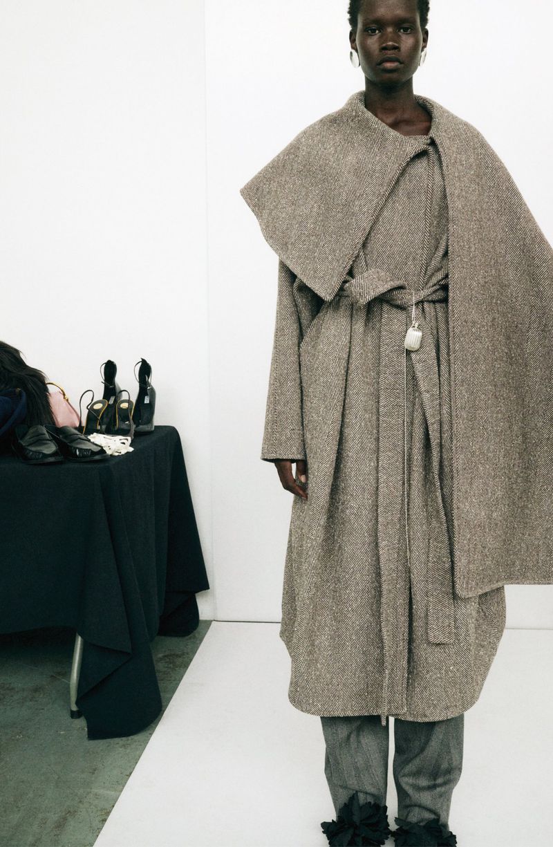 A tall, slender African-American woman poses in a neutral studio. She wears an oversized brown coat and gray pants with dark ruffles.