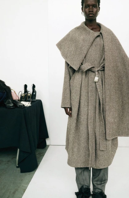 A tall, slender African-American woman poses in a neutral studio. She wears an oversized brown coat and gray pants with dark ruffles.