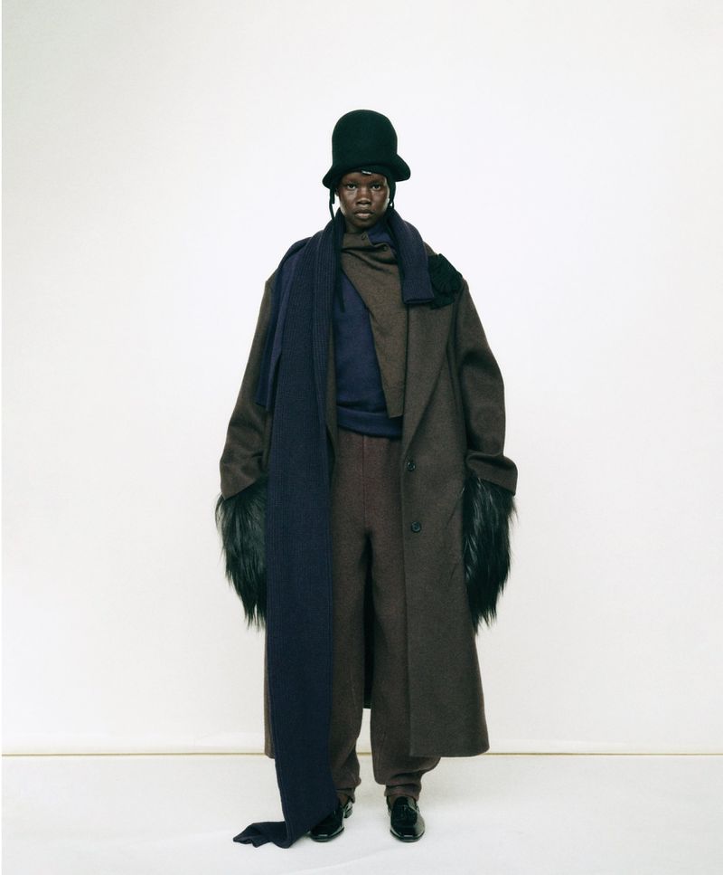 Full-length studio shot of adult model wearing a long dark brown coat with fur sleeves, dark blue scarf, dark brown pants, and a green hat.