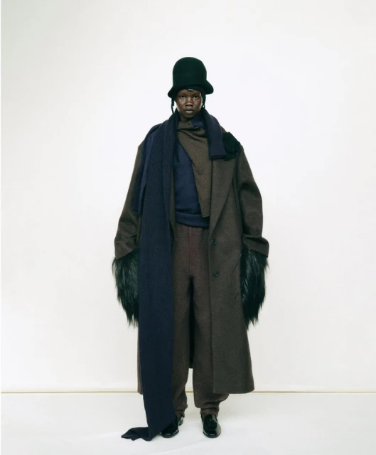 Full-length studio shot of adult model wearing a long dark brown coat with fur sleeves, dark blue scarf, dark brown pants, and a green hat.