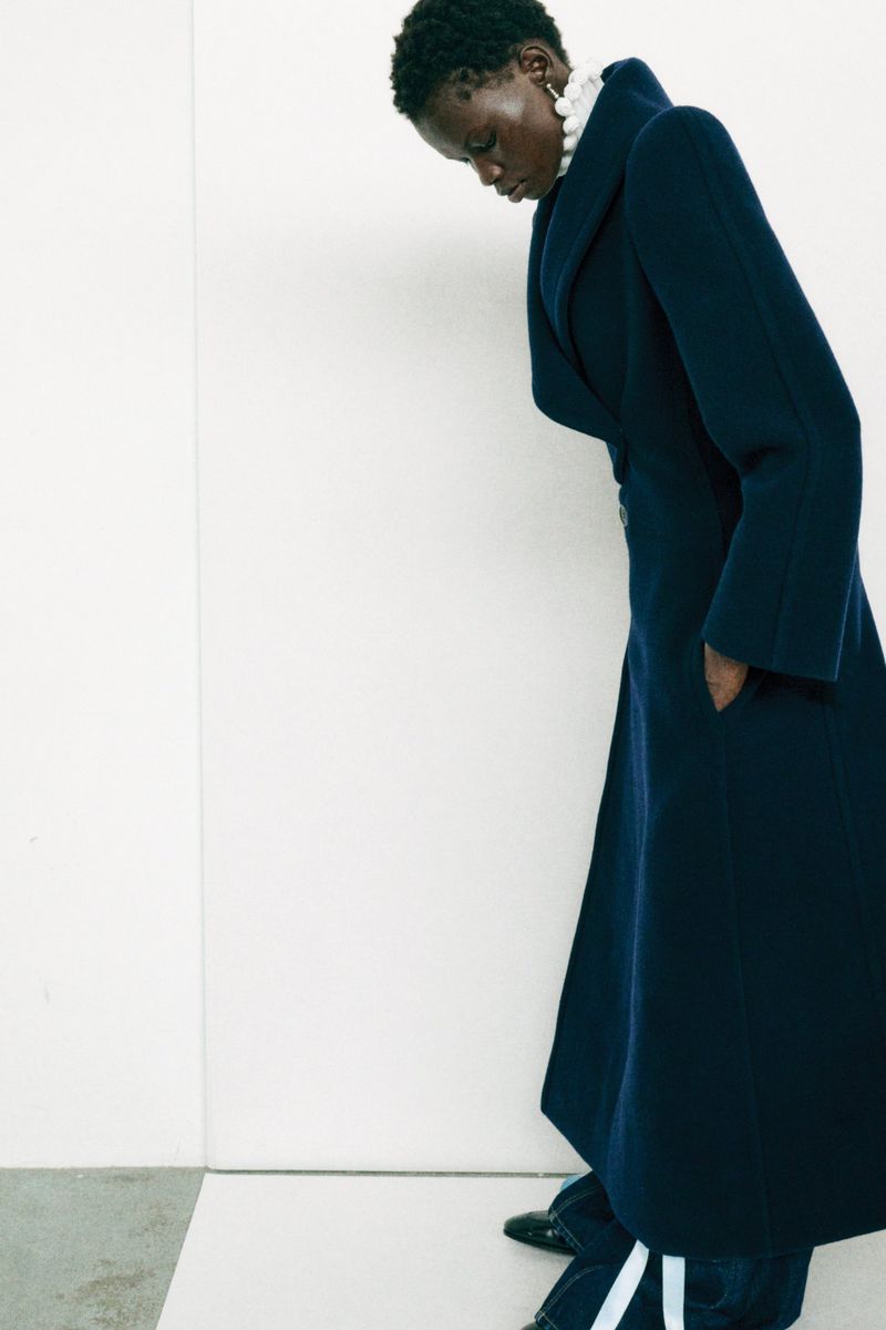 Dark-skinned woman wearing a dark blue wool coat with a wide collar and pearl earrings in a fashion shot against a bright white background.