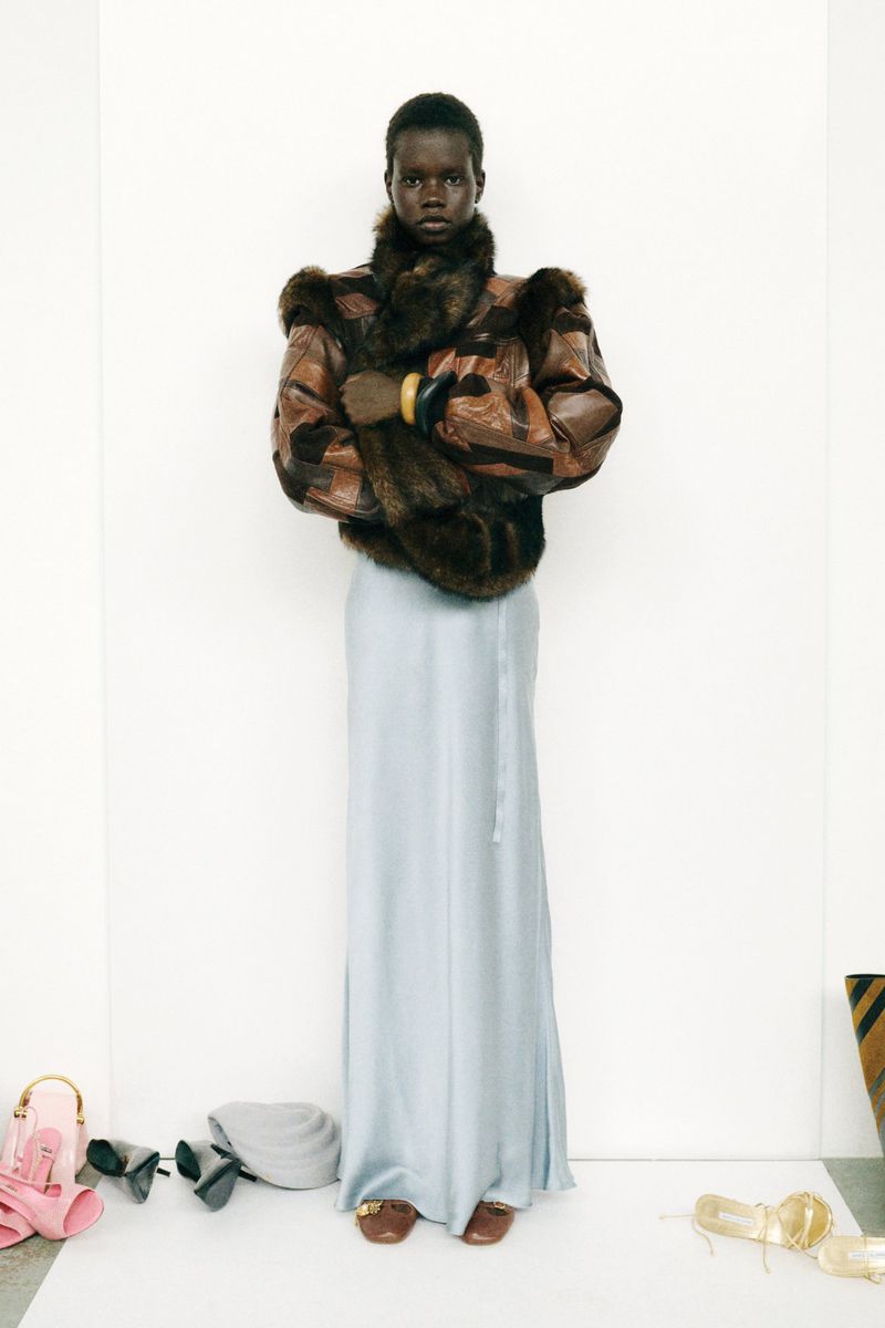 Elegant dark-skinned woman in a fur-trimmed jacket and flowing skirt, standing in front of a blank white wall, with scattered accessories.