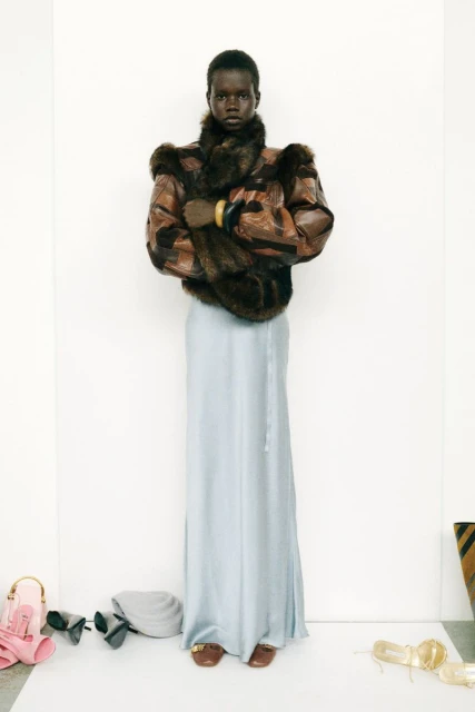Elegant dark-skinned woman in a fur-trimmed jacket and flowing skirt, standing in front of a blank white wall, with scattered accessories.