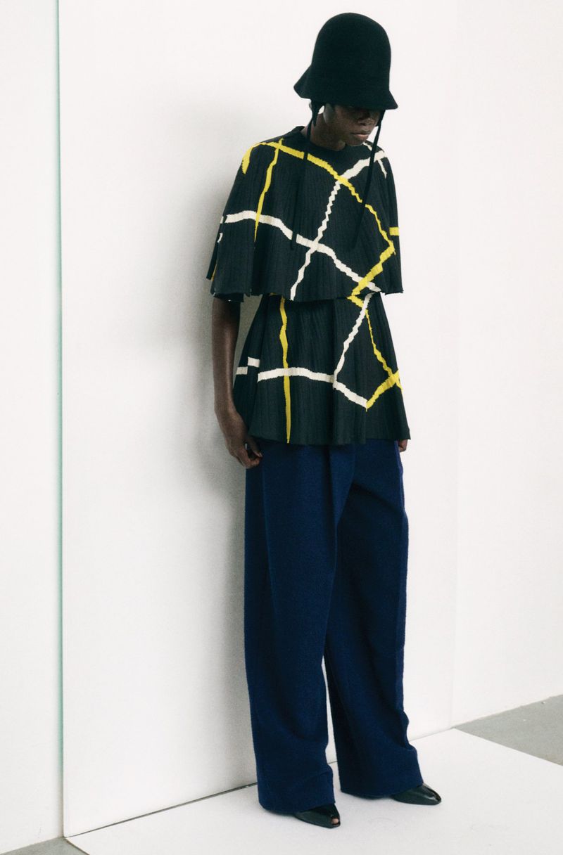 Fashion model wearing a black hat, top with yellow and white geometric pattern, and wide-leg dark blue pants in a studio setting.