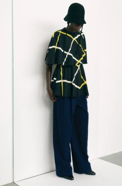 Fashion model wearing a black hat, top with yellow and white geometric pattern, and wide-leg dark blue pants in a studio setting.