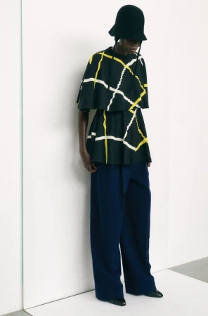 Fashion model wearing a black hat, top with yellow and white geometric pattern, and wide-leg dark blue pants in a studio setting.
