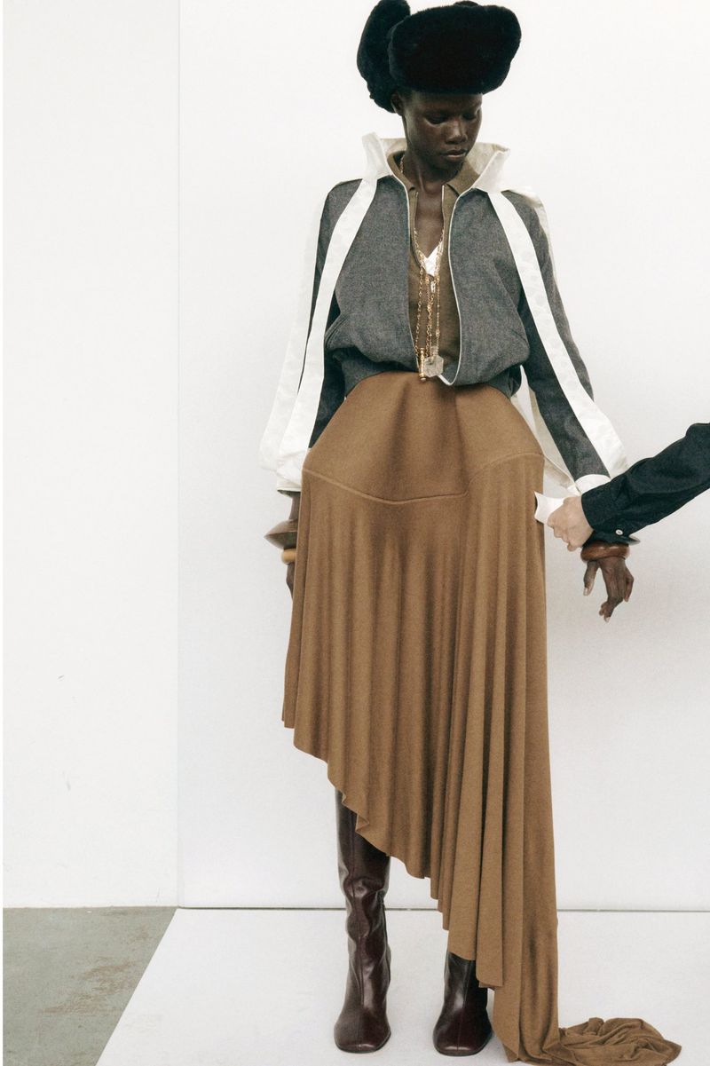 Runway fashion show features a Black model in a gray jacket, brown skirt, leather boots, and fur hat against a white background.