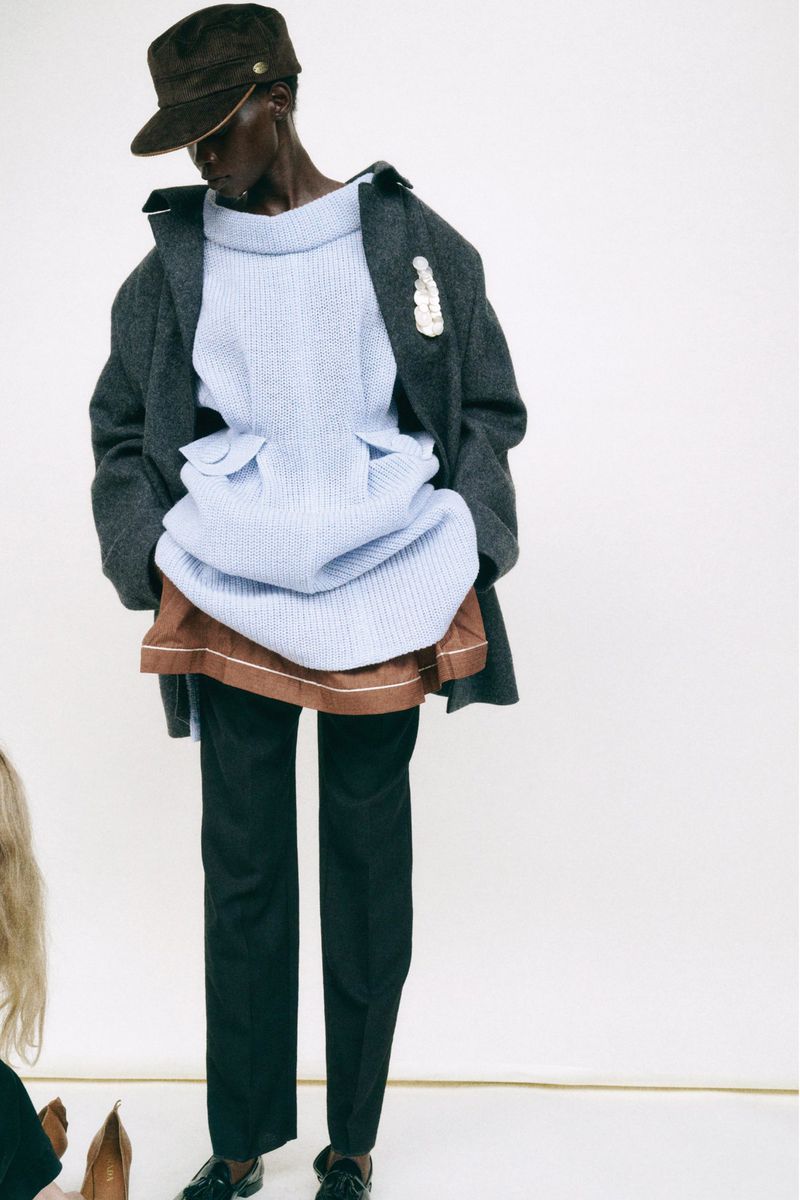 A tall fashion model wearing a blue sweater and black trousers poses in front of a white backdrop wearing a dark-grey coat and a hat.
