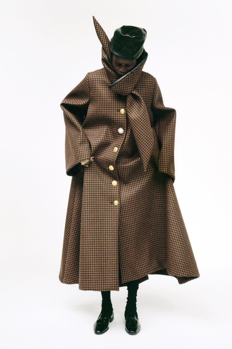 Fashionable adult in houndstooth coat and headwear stands in a photo studio with a neutral background, highlighting modern apparel and style.