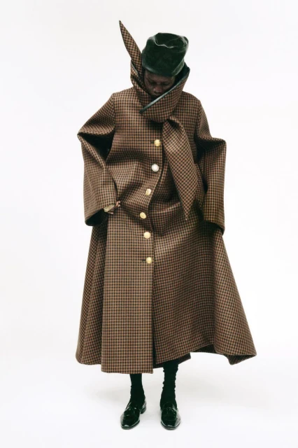 Fashionable adult in houndstooth coat and headwear stands in a photo studio with a neutral background, highlighting modern apparel and style.