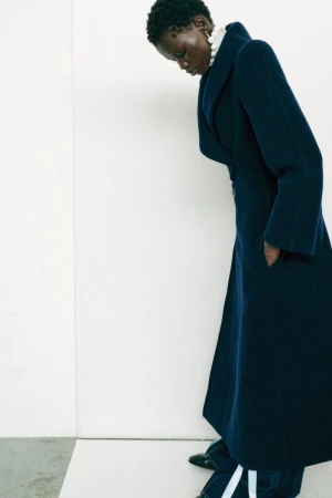 Model Mary Ukech in a long navy blue structured coat looking down against a plain white studio backdrop.