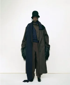 Model Mary Ukech standing in an oversized brown wool coat with fur cuffs, dark hat, and trailing navy scarf on a white background.