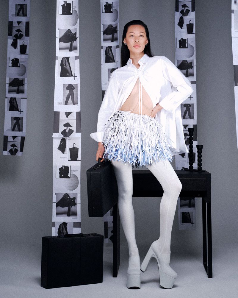 A fashion-forward Asian model in a white shirt, blue/silver fringed skirt, and platform heels poses with briefcases in a studio setting.